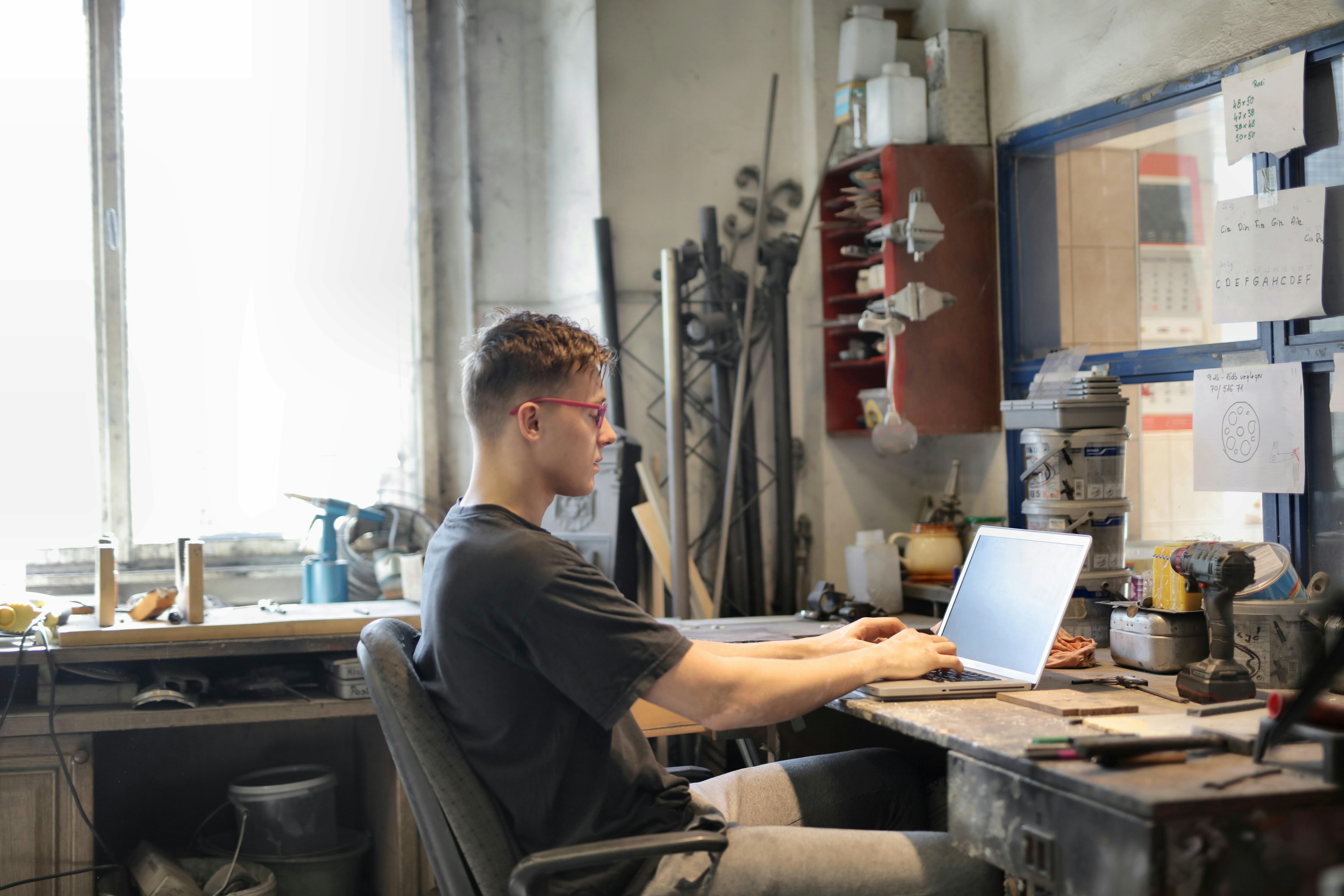 A young man works on a laptop in a creative workshop setting, surrounded by tools and materials.